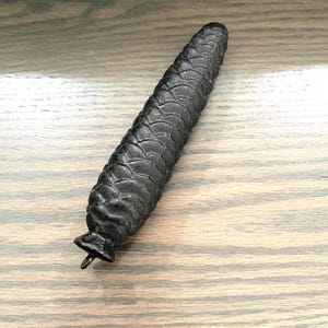 May include: A black, elongated, cylindrical weight with a textured, scale-like surface pattern resembling a pinecone. The object features a small, flared base with a metal loop extending from the bottom, designed for hanging or attachment purposes.
