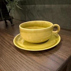 May include: A chartreuse ceramic teacup and matching saucer set. The teacup features a smooth, glossy finish with a rounded bowl shape and a small, curved handle on the side. The saucer is circular with a slightly raised edge, matching the cup.