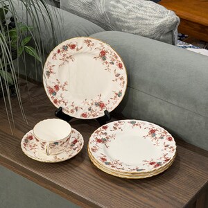 Könnte beinhalten: Ein cremefarbenes Porzellangeschirr-Set mit einem zarten Blumenmuster in Rot- und Blautönen sowie goldfarbenen, gewellten Rändern. Das Set besteht aus einer Teetasse auf einer passenden Untertasse, einem aufrechten Teller und zwei gestapelten Tellern.