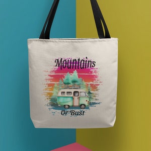 May include: A white canvas tote bag with black straps. The bag features a graphic of a vintage camper van with trees and a sunset in the background. The text "Mountains or Bust" is printed below the van.