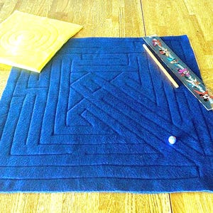 May include: A blue felt maze game with a square design, a yellow square, a ruler, a wooden stick, and a small marble. The maze has a geometric pattern. The yellow square has a spiral design.