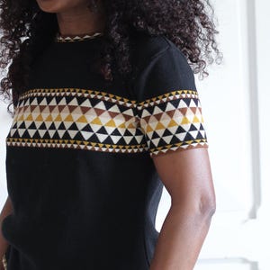 Peut inclure: Pull-over noir à manches courtes avec un motif géométrique dans les tons marron, jaune et blanc sur la poitrine et les manches. Le pull est associé à un jean bleu et une ceinture marron. La personne porte des lunettes de soleil.