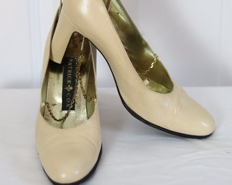Op de afbeelding: Een paar crèmekleurige leren pumps met een gouden kettingdetail. De schoenen zijn van het merk "Patrick Cox".