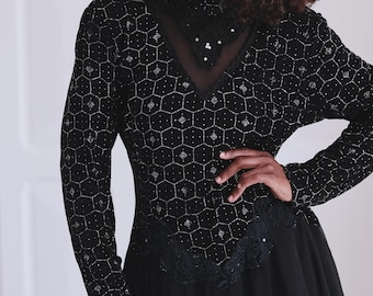 May include: Black long-sleeved dress with a geometric pattern of silver embellishments. The bodice features a V-neck and a sheer neckline. The skirt is a flowing, solid black fabric. The dress has a vintage aesthetic.