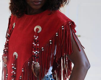 Può includere: Un poncho in camoscio rosso intenso con lunghe frange lungo i bordi. Il capo è decorato con conchos metallici circolari color argento, strisce di pelle pendenti ornate da piccole perline bianche e nere e piume marroni naturali.