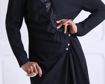 May include: A black long-sleeved dress with a ruffled detail on the left side. The dress has a V-neckline and a high-low hemline. The dress is cinched at the waist with a belt.