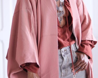 May include: A dusty rose coloured kimono with a silver chain necklace featuring large white stones. The kimono is worn over a rust coloured top and light grey jeans.