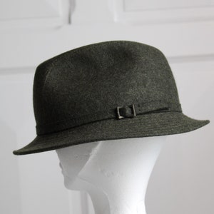 Peut inclure: Un chapeau fedora en feutre vert foncé avec un bandeau en cuir marron et une boucle argentée. Le chapeau est sur une tête de mannequin blanche.