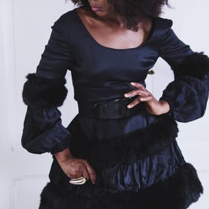 Op de afbeelding: Een zwarte satijnen jurk met een bontkraag en bontmanchetten. De jurk heeft een aansluitend bovenlijf en een volle rok. Het model draagt een zwarte riem met een grote gesp.