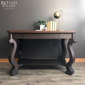 May include: A dark brown wooden desk with a single drawer and a lower shelf. The desk has ornate, curved legs and a gold lamp with a round shade on the top.