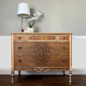 May include: A vintage wooden dresser with four drawers and a light wood finish. The dresser has brass hardware and sits on four turned legs with casters. The dresser is in front of a white wall with a white lamp and a plant on top.