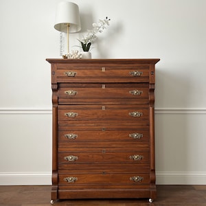 May include: A tall, antique wooden dresser with seven drawers and brass hardware. The dresser is a rich brown color and has a slightly distressed finish. It is sitting on a wooden floor in front of a white wall.