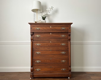 May include: A tall, antique wooden dresser with seven drawers and brass hardware. The dresser is a rich brown color and has a slightly distressed finish. It is sitting on a wooden floor in front of a white wall.