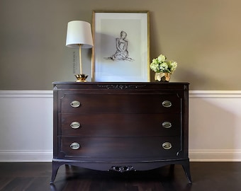 May include: A dark brown wooden dresser with three drawers and ornate detailing. The dresser has brass handles and sits on a dark wood floor. A gold lamp and a framed print are on top of the dresser.