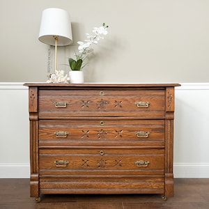May include: A vintage wooden dresser with three drawers and ornate carvings. The dresser has brass hardware and sits on small wheels. A white lamp with a white lampshade and a white vase with a white orchid are on top of the dresser.