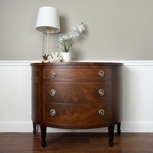 May include: A three-drawer, oval-shaped, wooden chest of drawers with brass hardware. The chest is a dark brown color and has a wood grain pattern. The chest is sitting on a wooden floor in front of a white wall.
