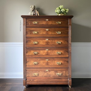 May include: A tall, antique wooden dresser with six drawers and brass hardware. The dresser has a rich, dark brown finish and is accented with a decorative trim around the top and bottom.