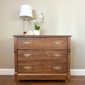May include: A vintage wooden dresser with three drawers and ornate brass hardware. The dresser is a rich brown color and has a slightly distressed finish. It is sitting on a wooden floor and has a white lamp and a potted plant on top.