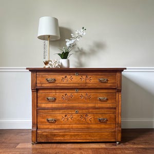 May include: A vintage wooden dresser with three drawers and carved floral details. The dresser is painted in a warm brown color and has brass hardware. It is sitting on a wooden floor in front of a white wall.