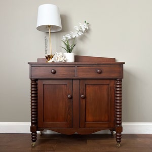 May include: A dark brown wooden cabinet with two drawers and two doors. The cabinet has turned legs and sits on small wheels. The cabinet is decorated with a white lamp, a vase of white flowers, and a gold decorative object.