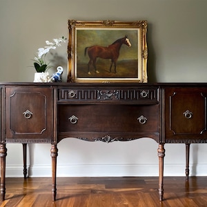 May include: A dark brown wooden buffet with ornate carvings and silver hardware. The buffet has two doors on the left, two drawers in the center, and two doors on the right. The buffet is sitting on a hardwood floor. A framed painting of a brown horse is hanging on the wall above the buffet.