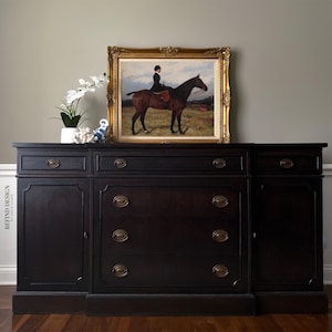 May include: A black wooden sideboard with four drawers and two cabinet doors. The sideboard has brass hardware and is decorated with a framed painting of a woman riding a horse.