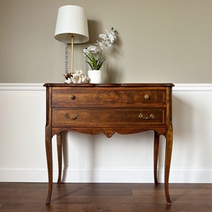 May include: A two-drawer wooden side table with a light brown finish. The table has a curved top and legs, and features ornate brass hardware. A white lamp with a white shade and a vase of white orchids are on top of the table.