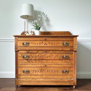 May include: A vintage wooden dresser with three drawers and ornate carved details. The dresser has brass hardware and is painted in a light wood stain. A white lamp and a white vase with white flowers are on top of the dresser.