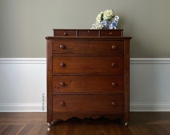 May include: A vintage wooden dresser with five drawers and a top shelf. The dresser is painted a rich brown color and has a slightly distressed finish. The dresser is on wheels and has a decorative trim around the bottom.