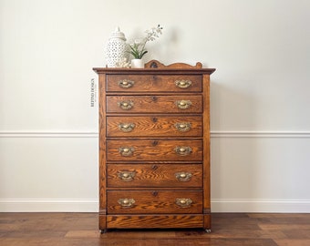 May include: A tall, antique wooden dresser with six drawers and brass hardware. The dresser is a warm brown color with visible wood grain. A decorative white ceramic jar and small plant sit on top. The words "REFIND DESIGN" are visible on the side.