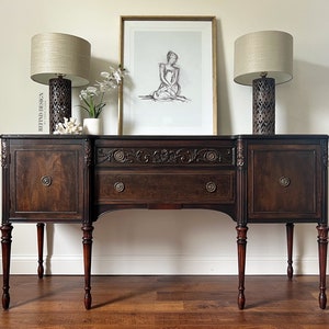 May include: A dark wood sideboard with intricate carvings and two doors on each side. The sideboard has a single drawer in the center and sits on four turned legs. Two lamps with beige shades and patterned bases are on the top of the sideboard, and a framed drawing of a woman is hanging on the wall behind it.