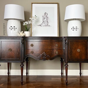 May include: A dark wood sideboard with three drawers and two cabinet doors. The sideboard has a curved front and sits on four turned legs. Two white ceramic lamps with white shades are on top of the sideboard, and a framed print of a woman is hanging on the wall behind the sideboard.