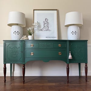 May include: A dark green buffet with carved floral details, brass hardware, and dark wood legs. Two white ceramic lamps with patterned bases flank a framed print. A small tassel hangs from the right door.