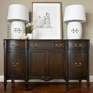 May include: A dark brown wooden buffet with three drawers and two doors. The buffet has ornate details and brass hardware. Two white ceramic lamps with white lampshades are on top of the buffet, along with a framed print and a potted plant.