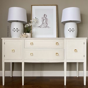 May include: White painted sideboard with two doors and two drawers. The sideboard has gold hardware and sits on four legs. Two white ceramic lamps with white shades are on top of the sideboard. A framed print of a woman sitting is above the sideboard.