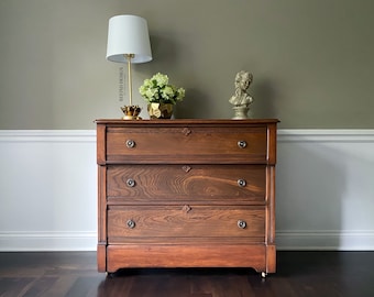 Op de afbeelding: Een dressoir met drie lades in een donkere houtkleur. Het dressoir heeft een eenvoudig ontwerp met messing beslag en staat op een donkere houten vloer.