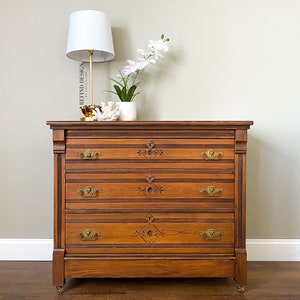 May include: A vintage wooden dresser with four drawers and brass hardware. The dresser has a dark wood stain and intricate carvings on the front of the drawers. The dresser is sitting on a wooden floor and has a white lamp and a white vase with white flowers on top of it.