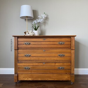 May include: A vintage wooden dresser with four drawers and brass hardware. The dresser is painted a warm honey brown color and has a slightly distressed finish. The dresser is sitting on a wooden floor and has a white lamp and a vase of white flowers on top.