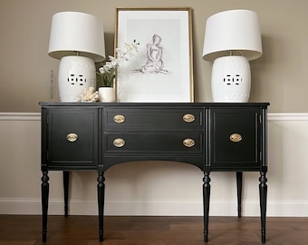 May include: Black painted sideboard with gold hardware and two drawers. The sideboard has four legs and is decorated with two white ceramic lamps with white shades.