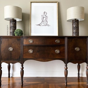 May include: A dark brown wooden sideboard with two doors on each side and a drawer in the center. The sideboard has turned legs and ornate brass hardware. Two lamps with beige shades and patterned bases are on top of the sideboard, and a framed drawing of a woman is hanging on the wall above the sideboard.