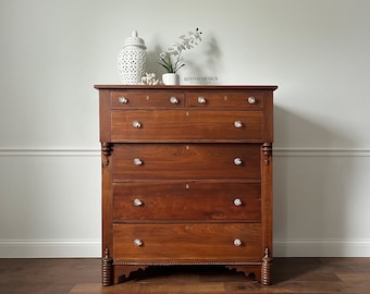 May include: A vintage wooden dresser with five drawers and crystal knobs. The dresser has a dark brown stain and intricate details on the bottom.