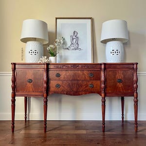 May include: A dark wood antique buffet with two white ceramic lamps and a framed sketch. The buffet has two cabinets, two drawers, and decorative carvings. The lamps have white shades and sit on white ceramic bases with cut-out designs.