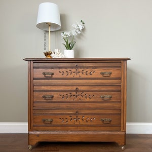 May include: A four-drawer wooden dresser with carved floral details and brass hardware. The dresser is painted a rich brown color and has a slightly distressed finish. It is sitting on a wooden floor with a white baseboard.