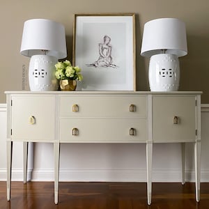 May include: A white painted sideboard with gold hardware and two drawers. Two white ceramic lamps with white shades are on top of the sideboard. A framed print of a woman sitting with a dog is on the wall above the sideboard.