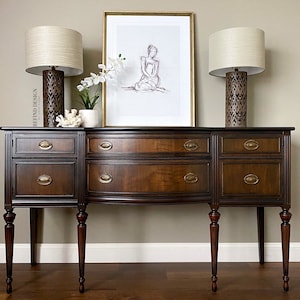 May include: A dark wood buffet with brass hardware, flanked by two lamps with beige shades. A framed sketch of a figure is centered above the buffet. Decorative items include a white coral piece and a potted plant.