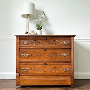 May include: A vintage wooden dresser with three drawers and intricate carved details. The dresser has brass hardware and a warm brown finish.