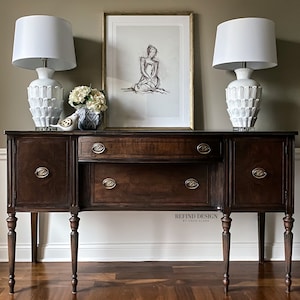 May include: A dark brown wooden sideboard with two doors on each side and a center drawer. The sideboard has four legs and brass hardware. Two white ceramic lamps with white lampshades are on top of the sideboard. A framed black and white drawing of a woman is hanging on the wall behind the sideboard.