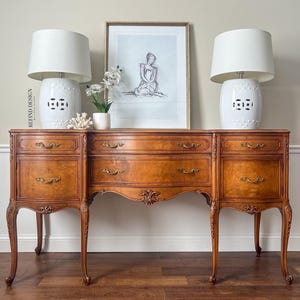May include: A vintage wooden buffet with a curved front and ornate details. The buffet has six drawers and is finished in a warm wood tone. Two white ceramic lamps with black accents are on top of the buffet, and a framed print of a woman is hanging on the wall behind it.