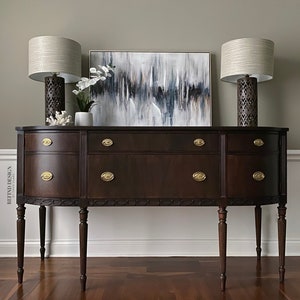 May include: A dark brown wooden sideboard with brass hardware and four drawers. Two lamps with beige shades and decorative bases are on top of the sideboard, along with a white vase with white flowers. An abstract painting with shades of blue, gray, and white hangs on the wall behind the sideboard.