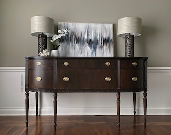 May include: A dark brown wooden sideboard with brass hardware and four drawers. Two lamps with beige shades and decorative bases are on top of the sideboard, along with a white vase with white flowers. An abstract painting with shades of blue, gray, and white hangs on the wall behind the sideboard.